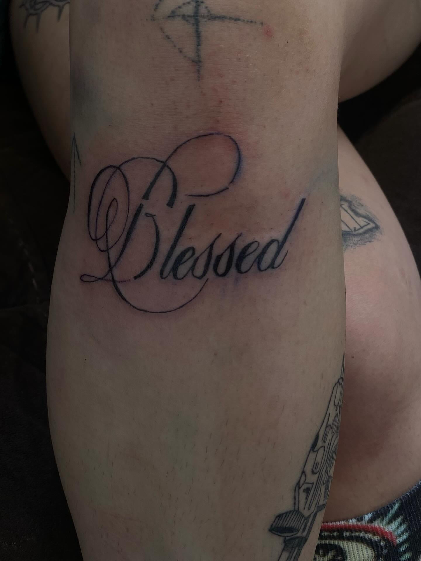 Blessed script lettering on the calf among earlier tattoos